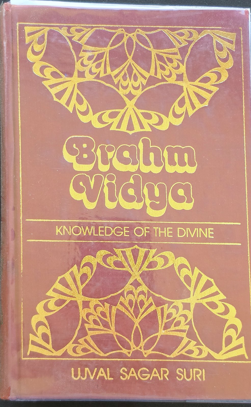 Maroon cover of Brahm Vidya: Knowledge of the Divine by Ujval Sagar Suri, with gold lotus designs, in a clear protective jacket
