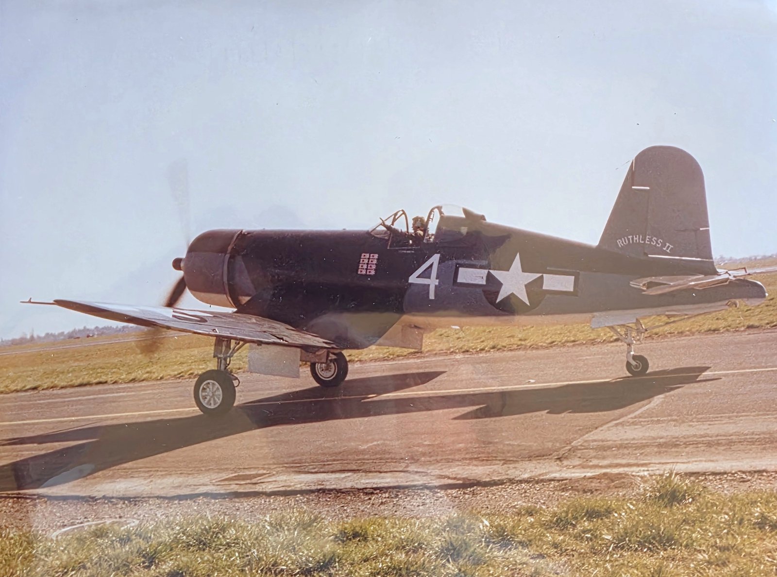 Color photograph of a dark blue F4U Corsair fighter with the number 4 and a star-and-bar insignia on the fuselage and the name Ruthless II on the tail, taxiing on a runway